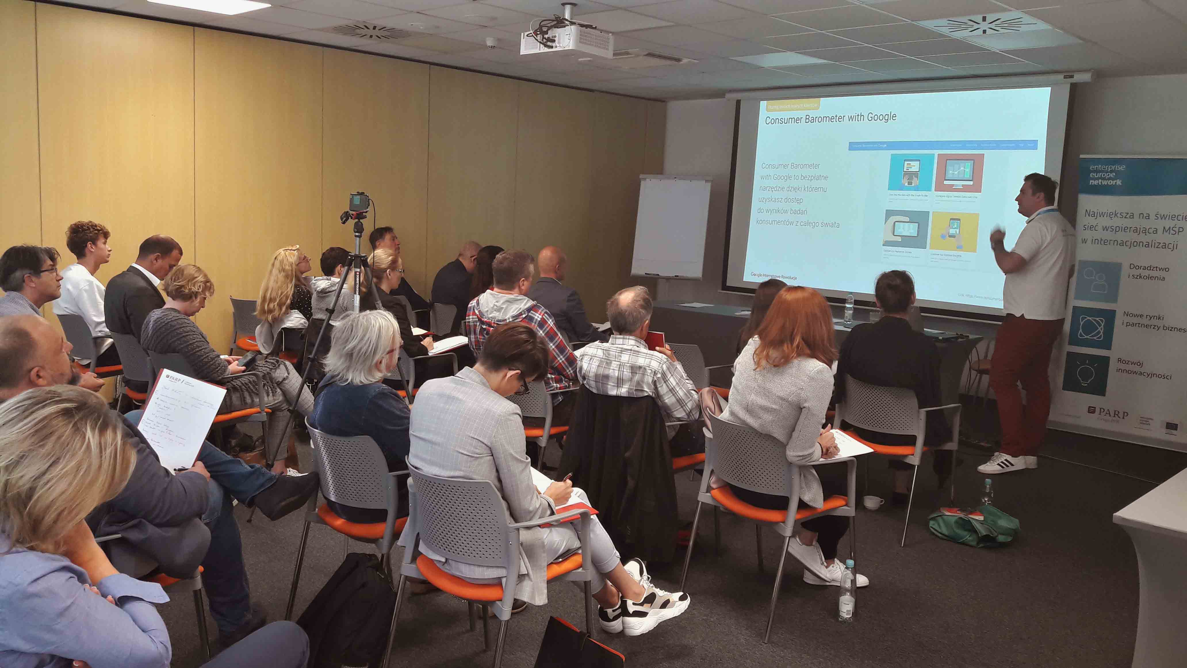 Sala wykładowa. Prowadzący przy ekranie mówi do siedzących uczestników