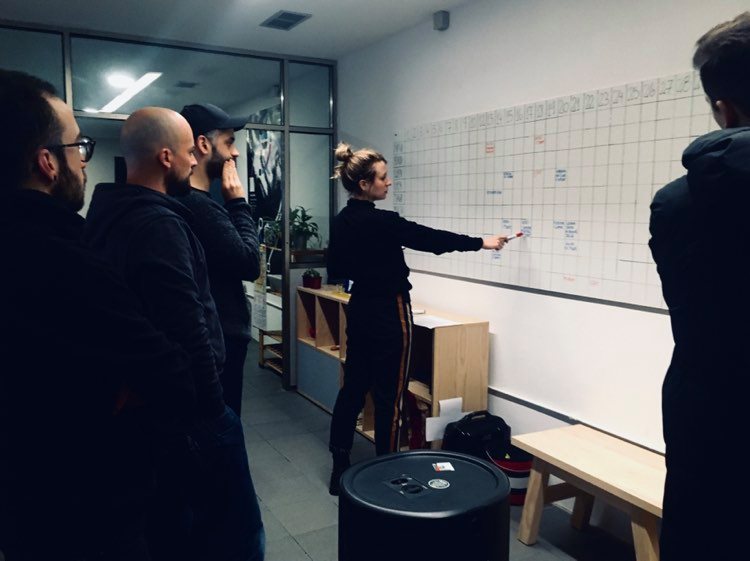 młoda kobieta ubrana w dres stoi przy białej tablicy z tabelkami i wskazuje na jedną z nich trzymanym w ręku flamastrem. Za kobietą stoi trzech mężczyzn w młodym i średnim wieku. Pierwszy z nich ma brodę i czapkę z daszkiem, drugi jest łysy i ma brodę, a trzeci ma kr&oacute;tkie czarne włosy i nosi okulary