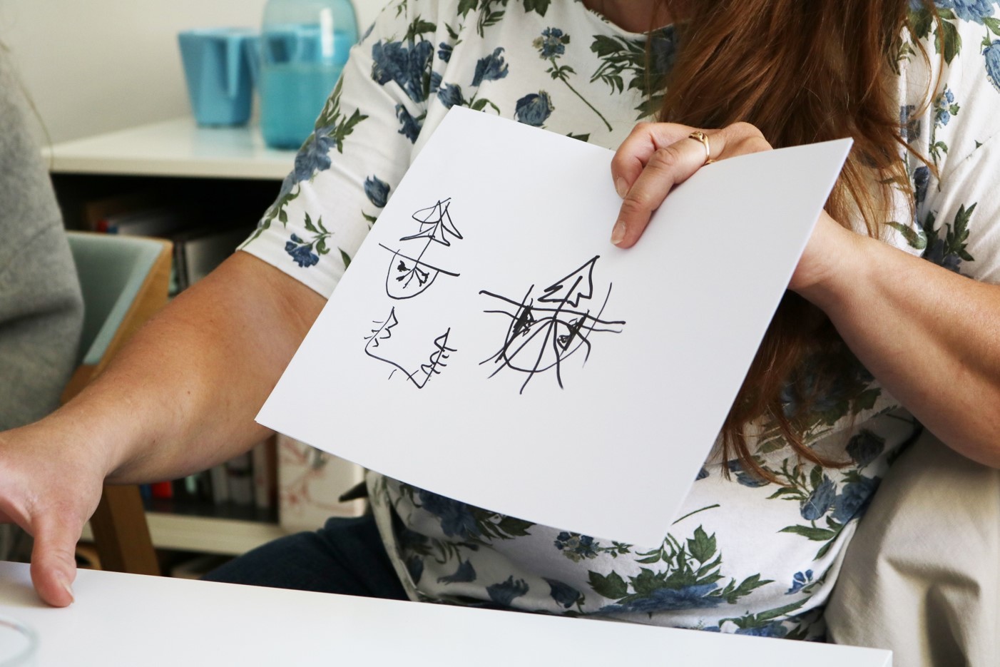 zbliżenie na ręce kobiety, kt&oacute;ra trzyma kartkę. Na białej kartce widać naniesiony czarny szkic, kt&oacute;re wyglądem przypomina doniczkę wkopaną w ziemię