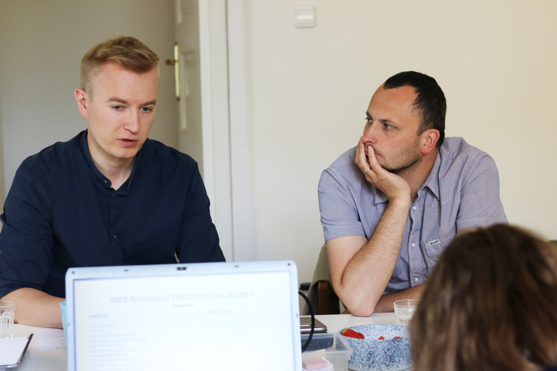 zbliżenie na dw&oacute;ch młodych mężczyzn siedzących przy stole. Mężczyzna widoczny po lewej stronie patrzy przed siebie, natomiast drugi z mężczyzn patrzy w jego kierunku. Na stole widać komputer typu laptop z otwartym ekranem