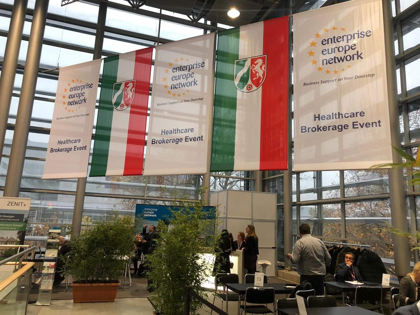 widok na przestrzeń do spotkań w dużym, mocno przeszklonym budynku z wysokim stropem. Pod sufitem są zawieszone banery, na kt&oacute;rych można przeczytać m.in. napis Enterprise Europe Network