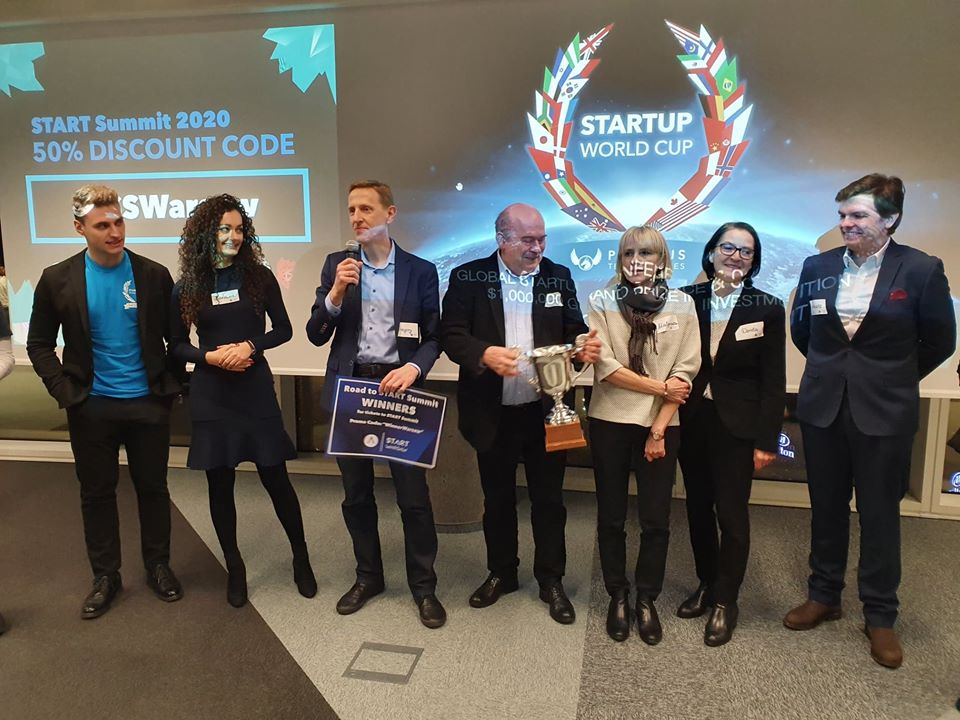 na zdjęciu widać siedem os&oacute;b, trzy kobiet i czterech mężczyzn, w r&oacute;żnym wieku, stojących w sztucznie oświetlonym pomieszczeniu. Za nimi widać ekran z angielskojęzycznym napisem  Startup World Cup. Jedna z os&oacute;b trzyma w ręku puchar