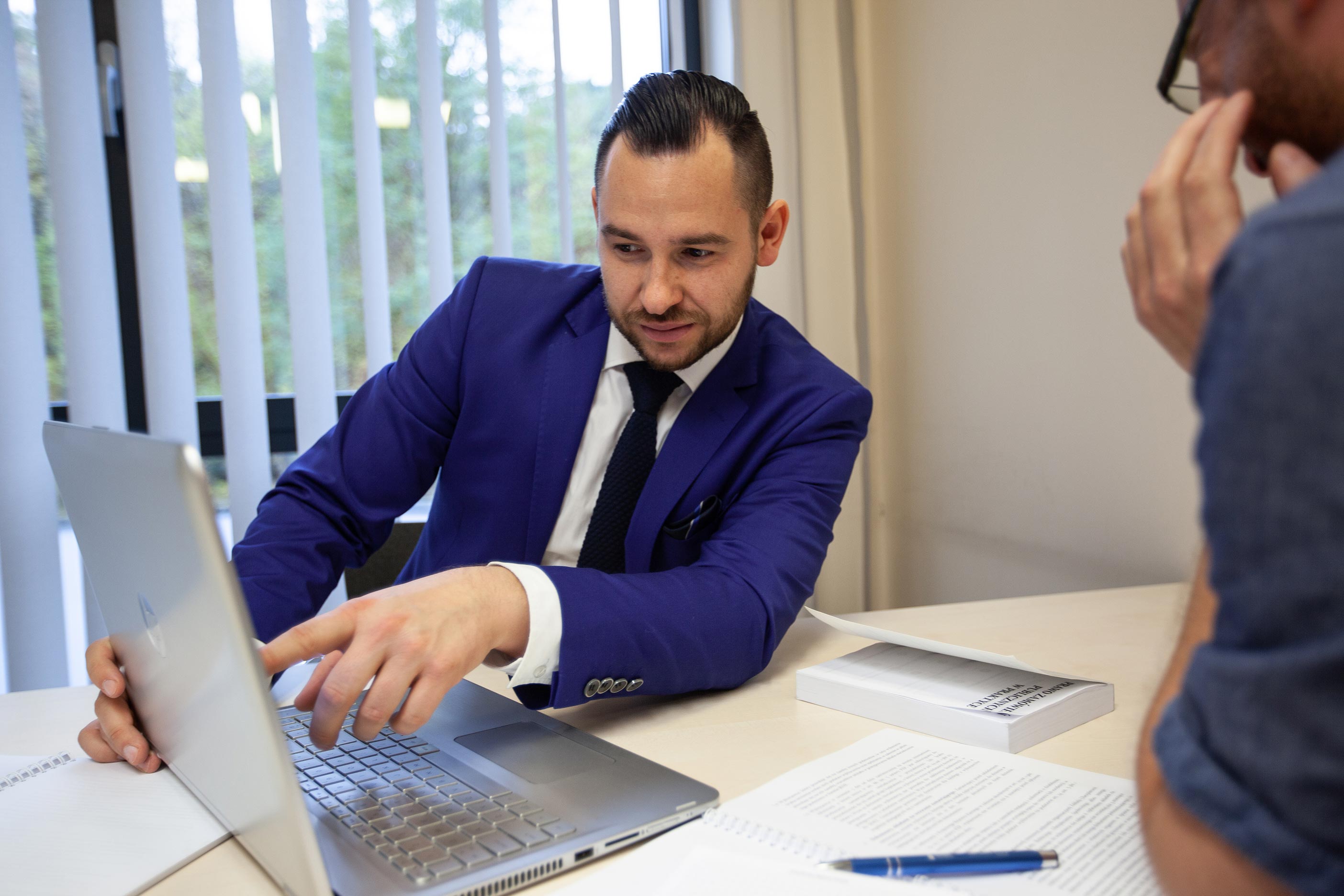 Widok dw&oacute;ch mężczyzn przy biurku, przybliżenie osoby w garniturze, kt&oacute;ra pokazuje coś ręką na ekranie komputera